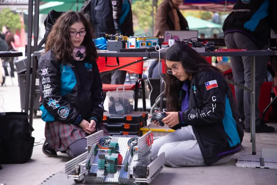 Two students in jackets work on a robotics project outdoors, assembling a wheeled metal chassis with circuit boards and tools nearby.