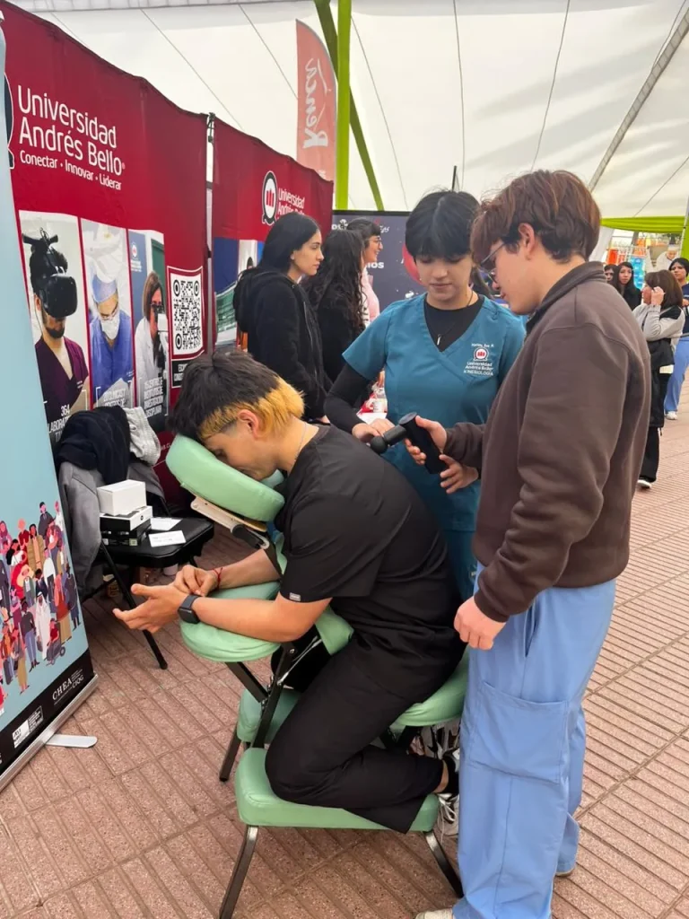 Man in black shirt sits in a green massage chair, resting his chin on a support while a healthcare worker in teal scrubs administers a neck massage at a university fair booth nearby people watch.