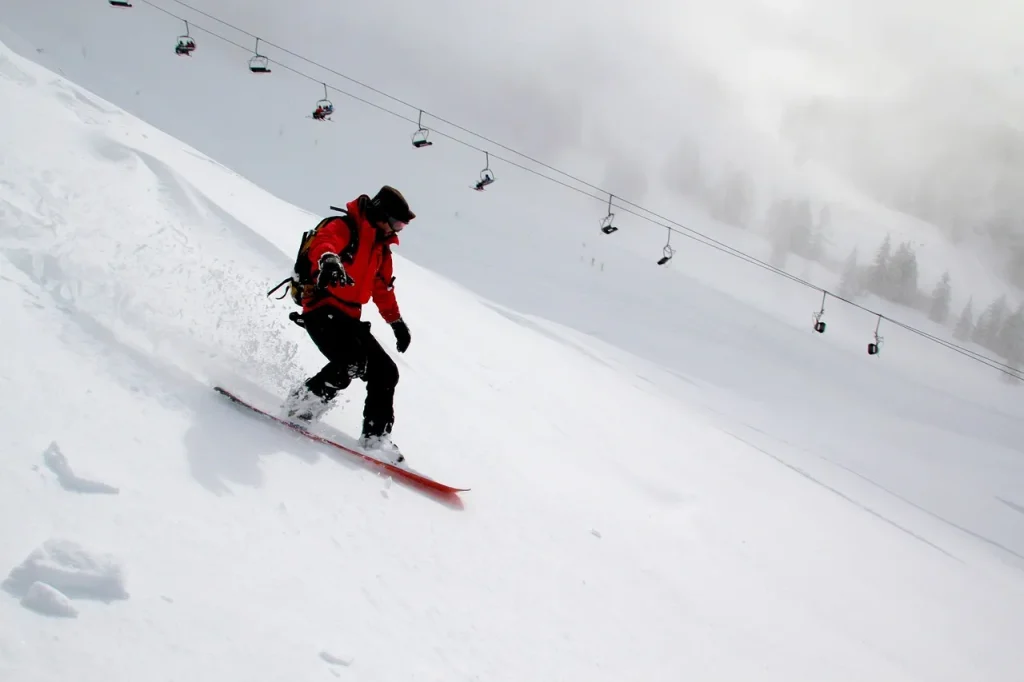 Skier in a red jacket carving down a snowy slope with a chairlift in the background.