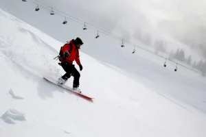 Skier in a red jacket carving down a snowy slope with a chairlift in the background.