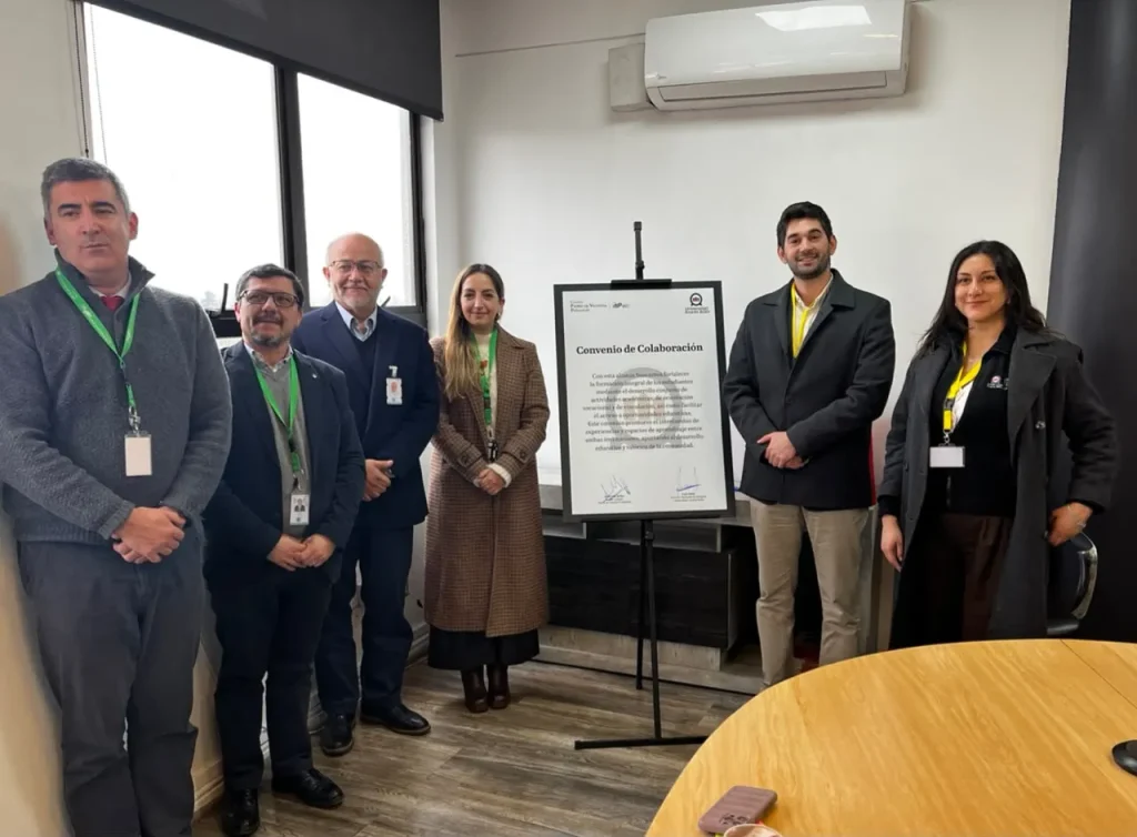 Six professionals in business attire pose in a conference room beside a framed 'Convenio de Colaboración' poster on a stand.