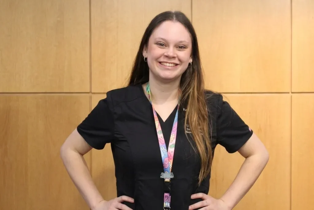 Smiling woman in black scrubs with a colorful lanyard and badge, standing with hands on hips in front of wood-paneled wall.