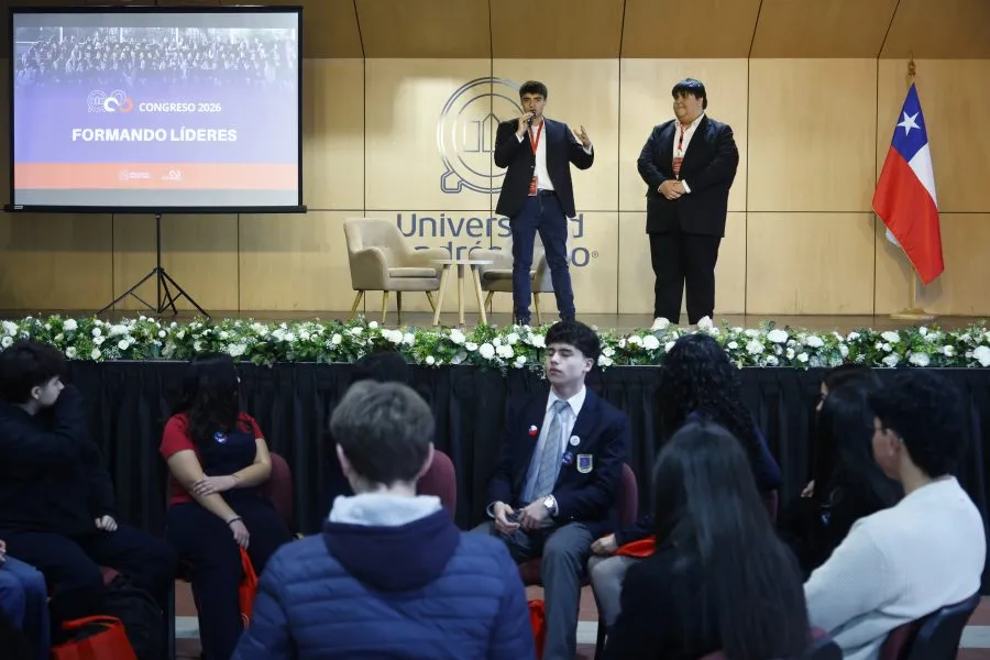 Two men on a stage giving a talk at a university conference; a Chilean flag hangs to the right and an audience sits facing them.