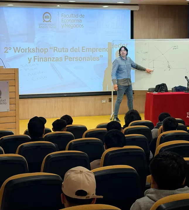 Male presenter in blue shirt points to a whiteboard during a lecture in a university auditorium; slide reads '2º Workshop Ruta del Emprendimiento y Finanzas Personales'.