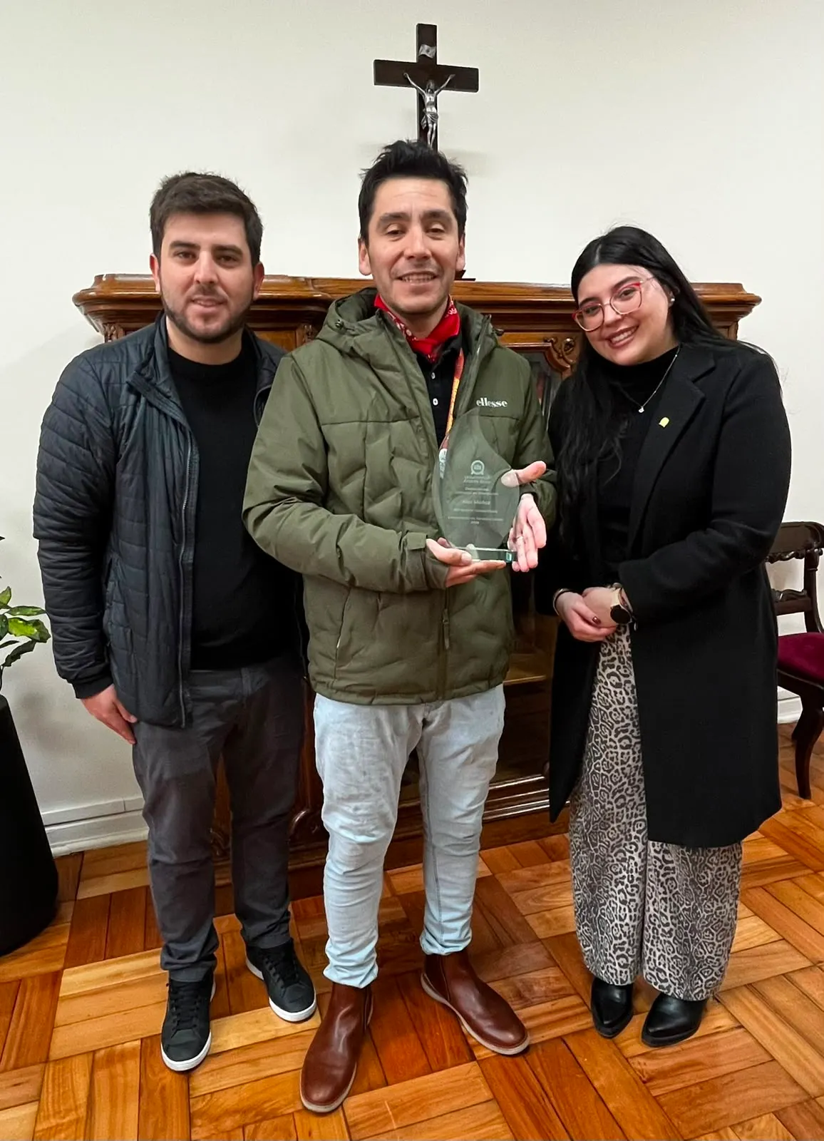 Three adults posing indoors; center man in green jacket holds a glass award, with a crucifix and wooden cabinet in the background.